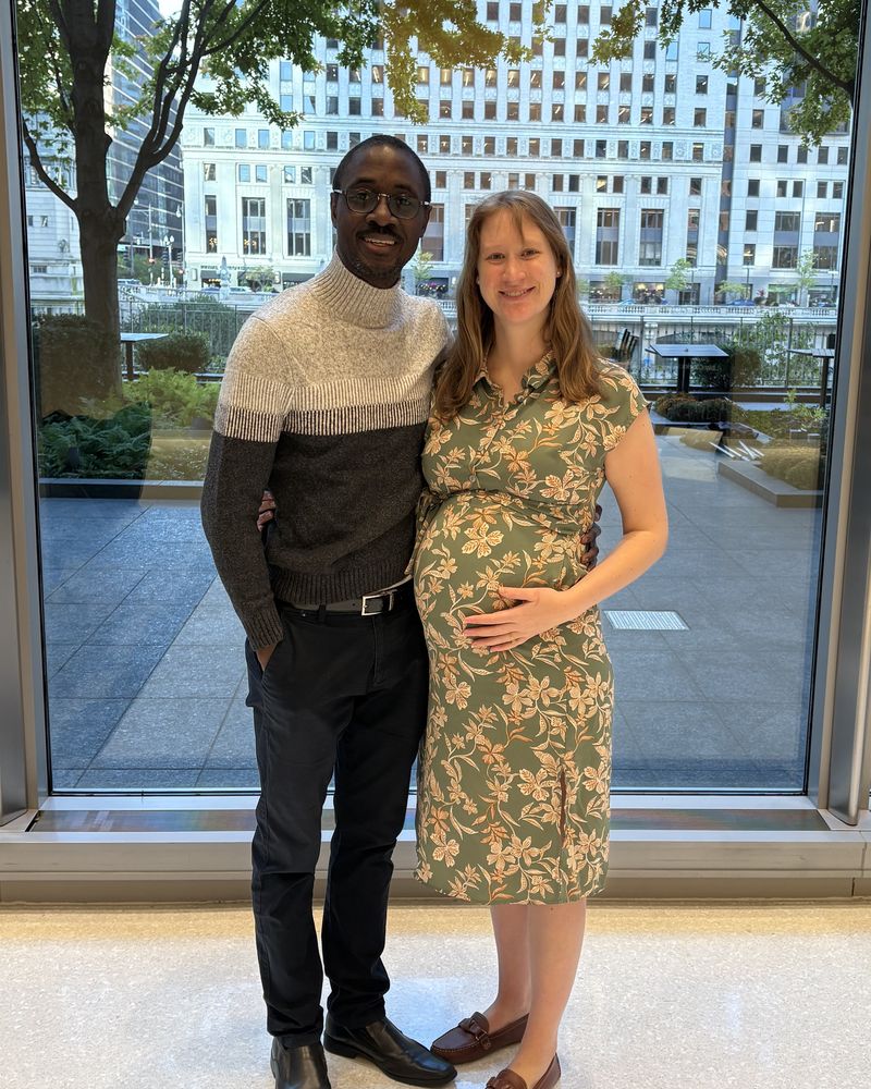 Daniel and Taylor smiling in front of a window overlooking the Chicago River; Taylor is cradling her baby bump.